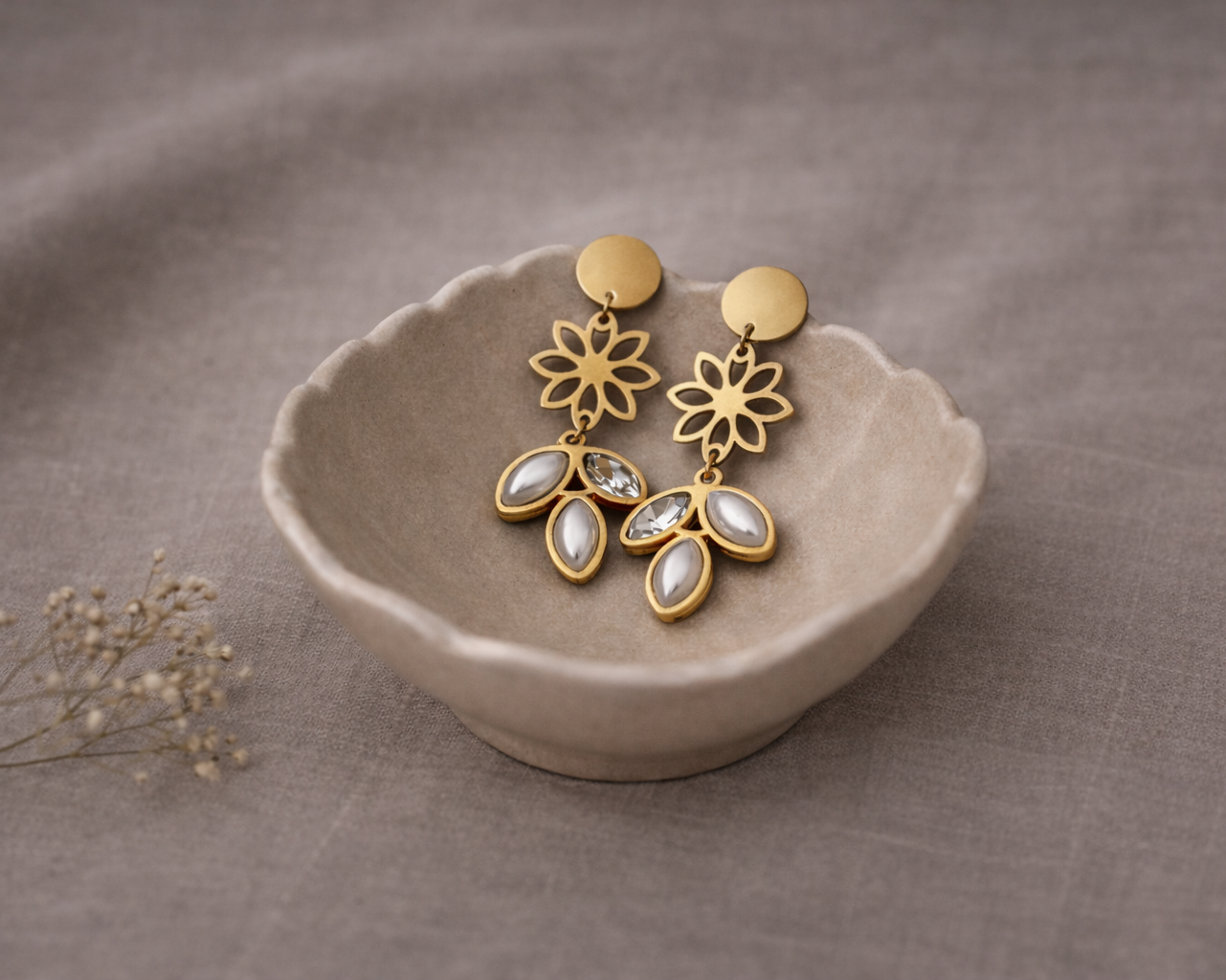 Gold floral earrings with clear stones on a beige dish against a brown fabric background