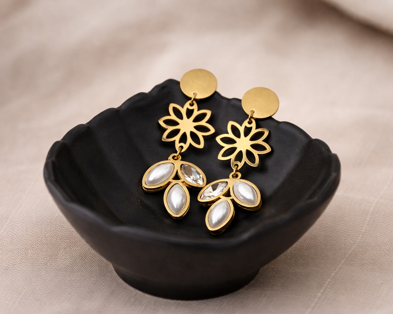 Gold and white floral earrings on a black dish with a beige background