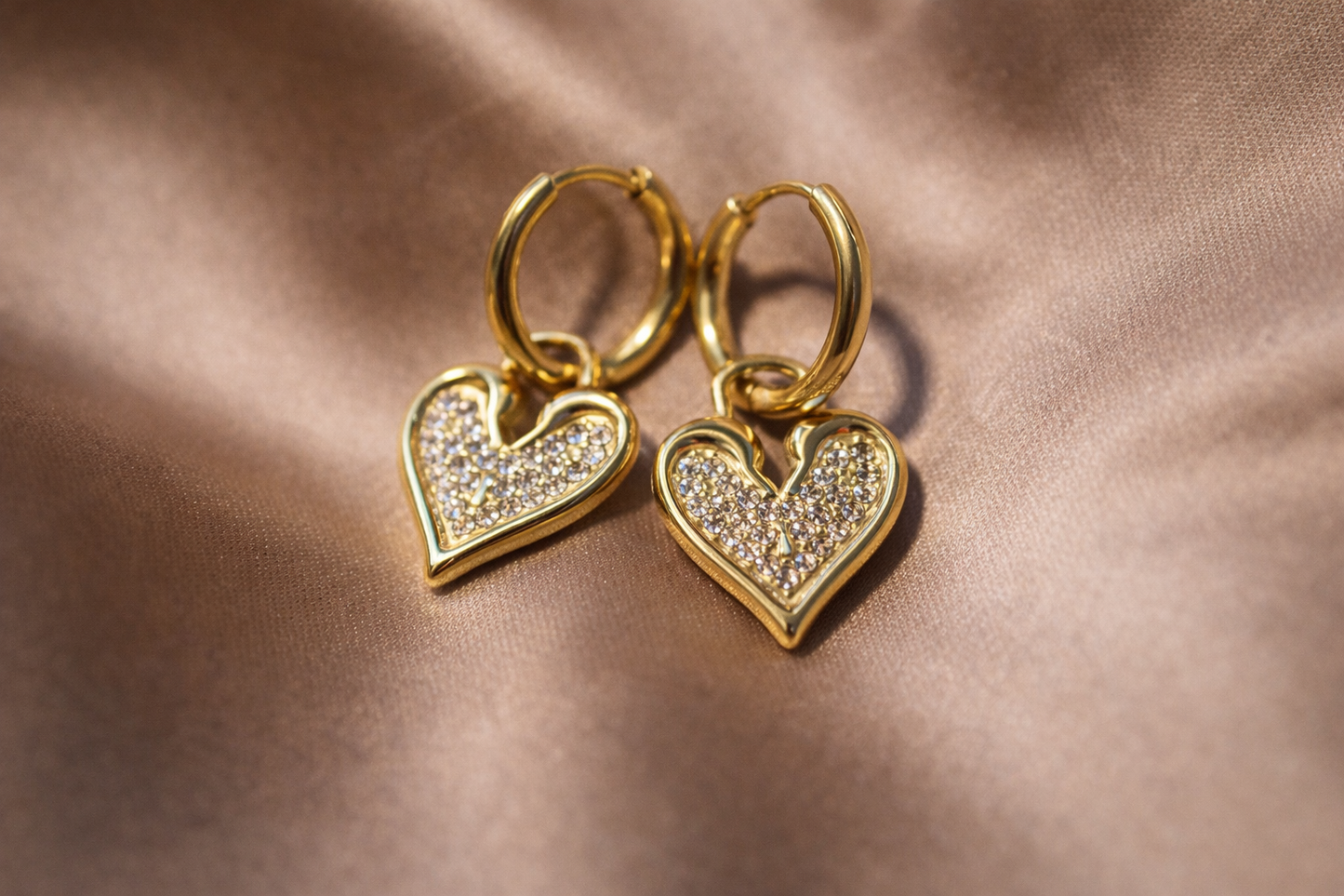 Gold heart-shaped earrings with diamond-like stones on a brown fabric background