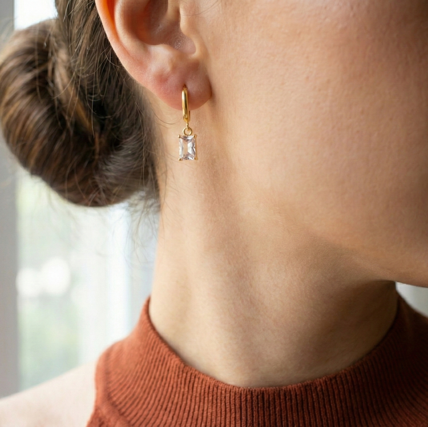 Close-up of a person wearing a gold earring with a diamond, set against a blurred indoor background.
